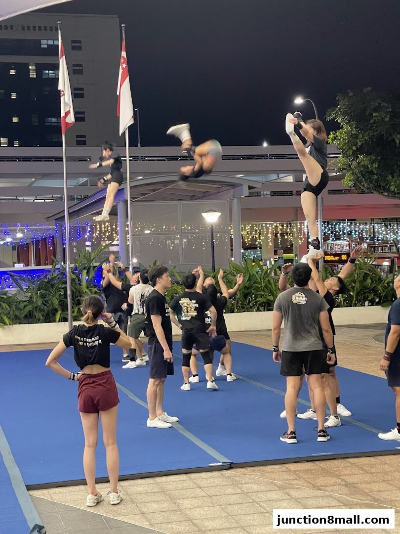 A cheerleading team practices stunts and tumbling on blue mats outdoors at night.