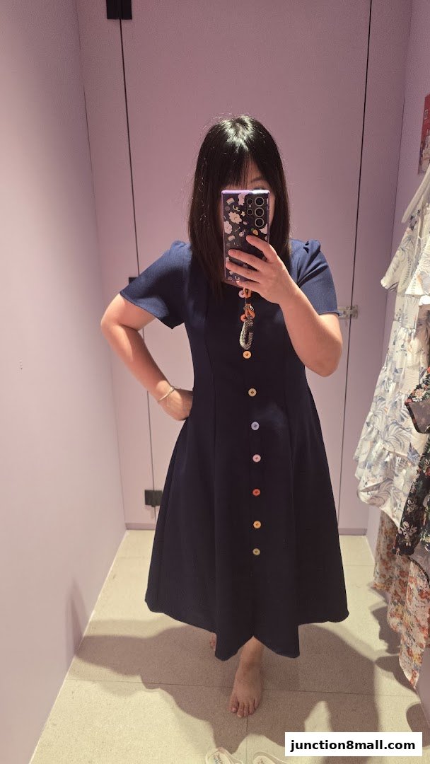 A woman tries on a navy blue dress with colorful buttons in a fitting room.