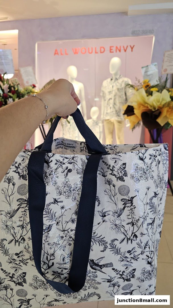 A person holds a patterned tote bag in front of the ALL WOULD ENVY store display.
