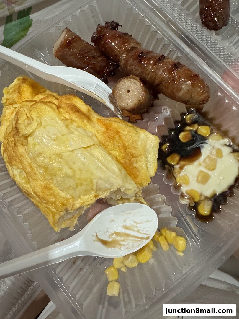 A close-up of a takeaway container with sausages, an omelet, and corn with sauce.