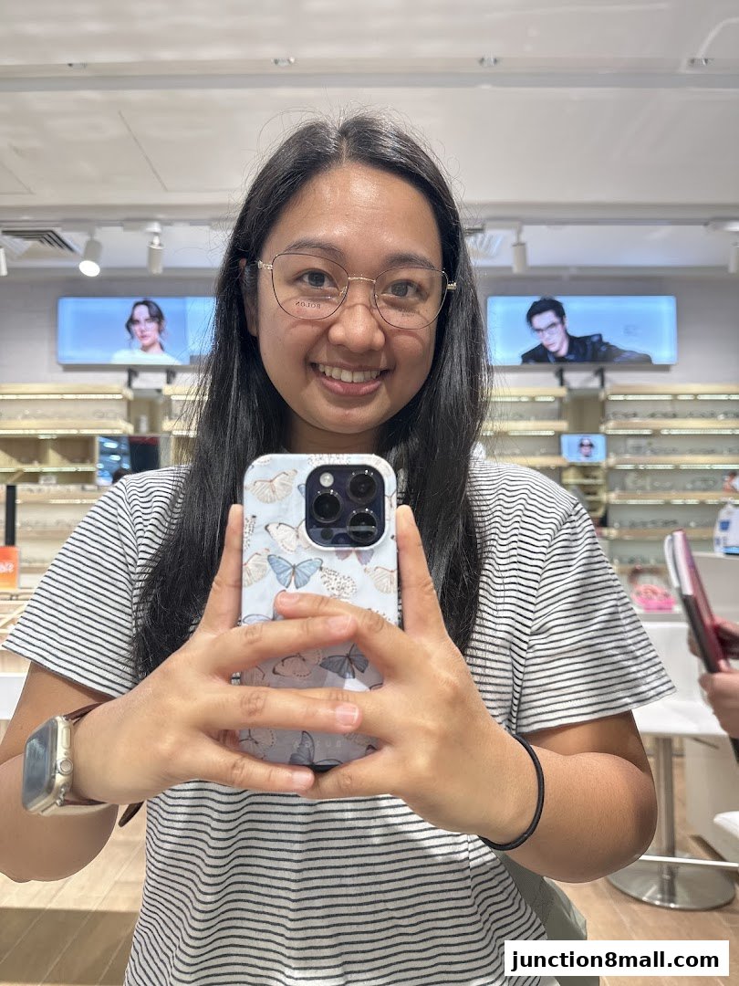 A woman smiles at the camera, wearing glasses and holding a phone in a Bolon Eyewear store.