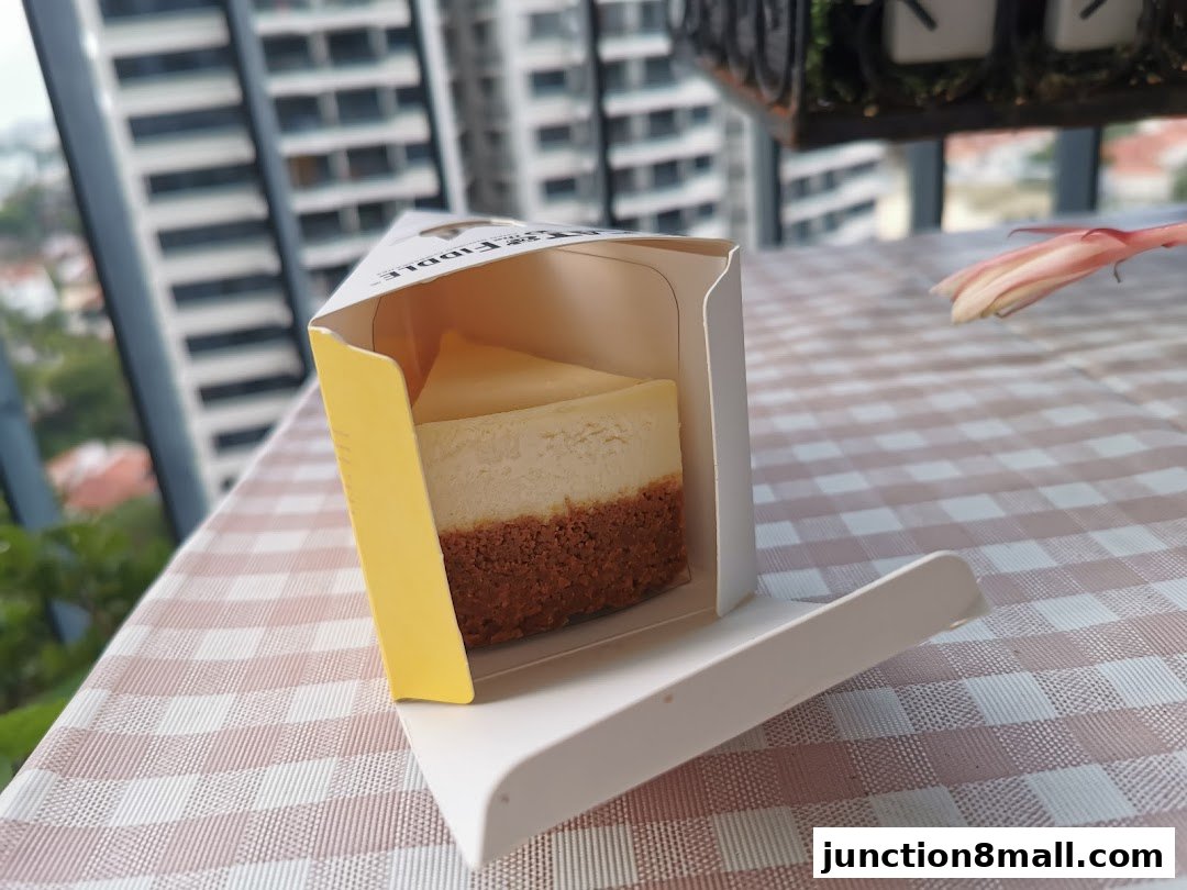 A slice of cheesecake in a branded box on a checkered tablecloth with a flower.