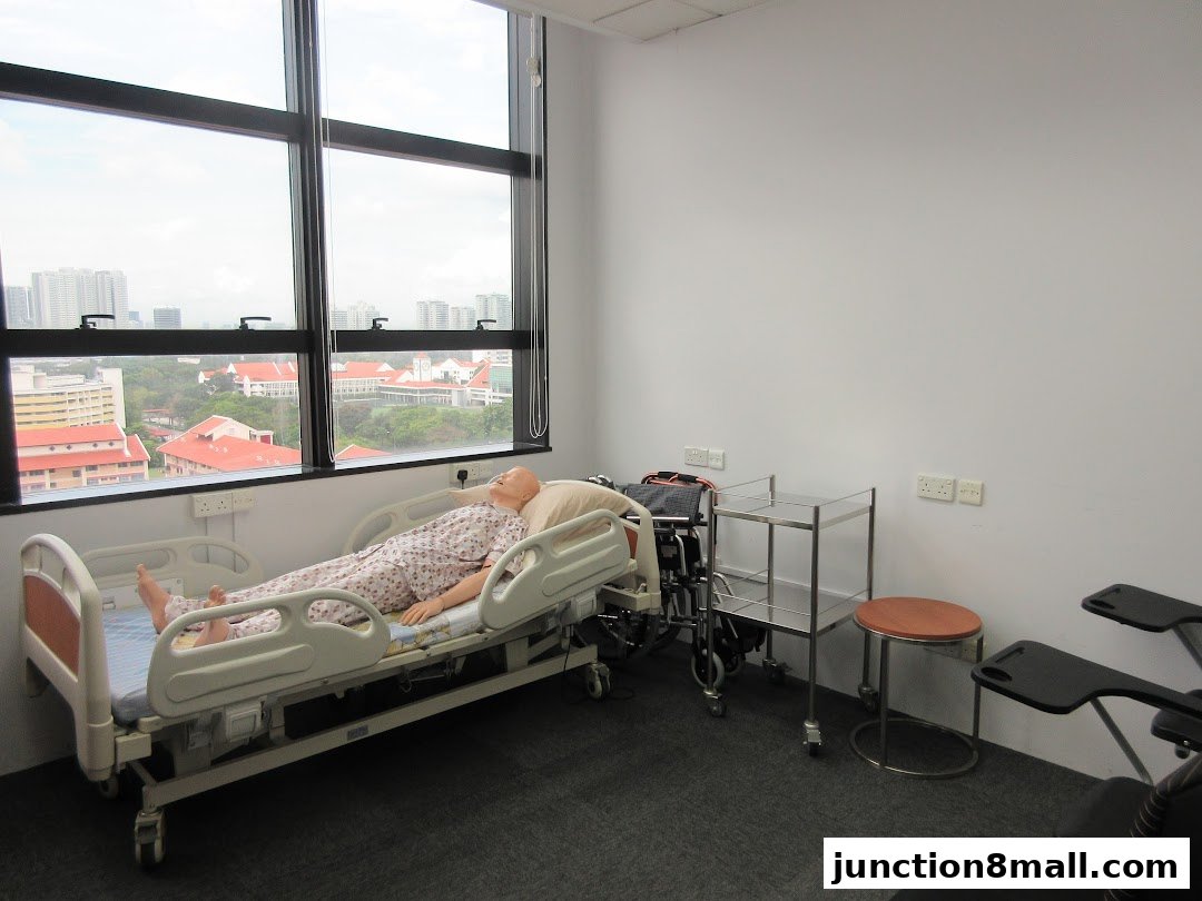 A medical training room with a patient simulator on a hospital bed and a wheelchair.