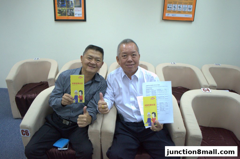 Two smiling men hold brochures in front of chairs at the Centre for Seniors.