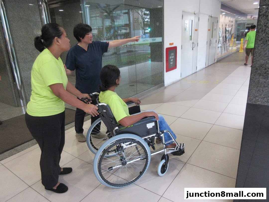 Two women assist a person in a wheelchair in a mall hallway.
