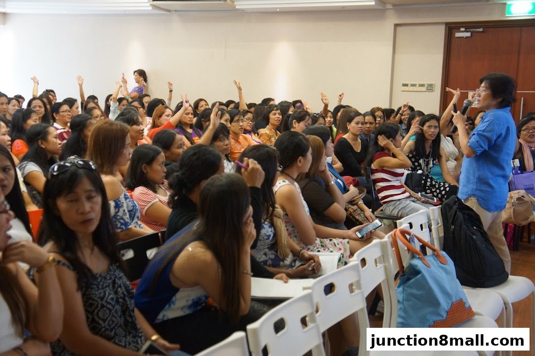 A speaker addresses a seated audience, many with hands raised, in a conference or seminar setting.