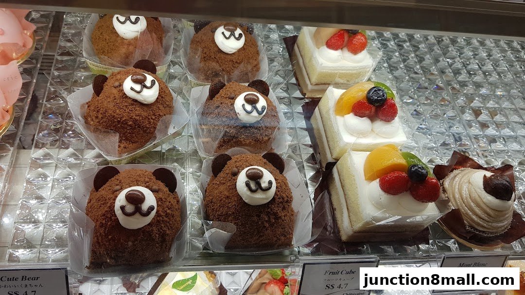 A display case filled with various cakes, including cute bear-shaped chocolate cakes and fruit-topped square cakes.