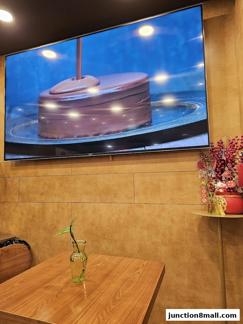 A television screen displays chocolate being poured onto a cake, with a table and vase in the foreground.