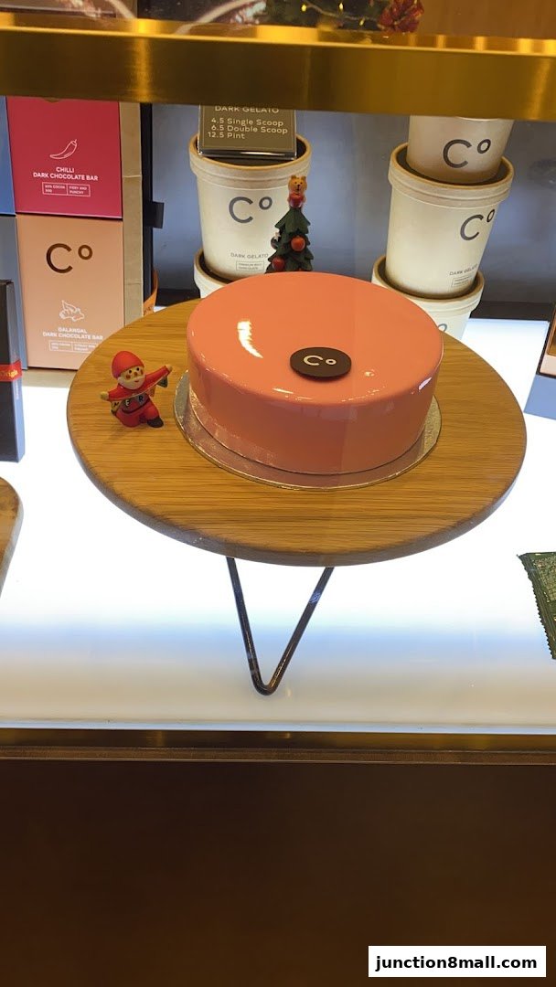 A glossy pink cake with a chocolate logo sits on a wooden stand, with gelato cups and chocolate bars in the background.