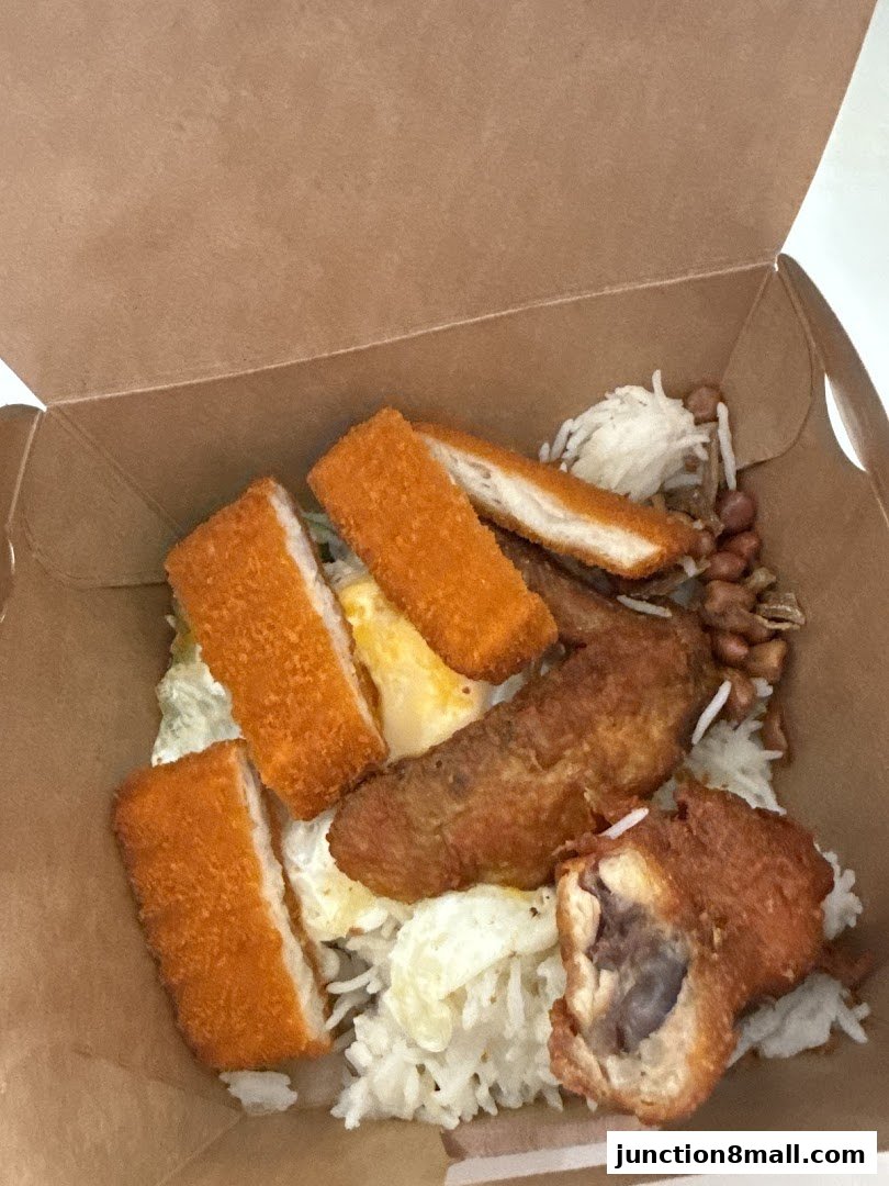 A close-up view of a takeaway box filled with Nasi Lemak, featuring fried chicken, fish fillet, and rice.