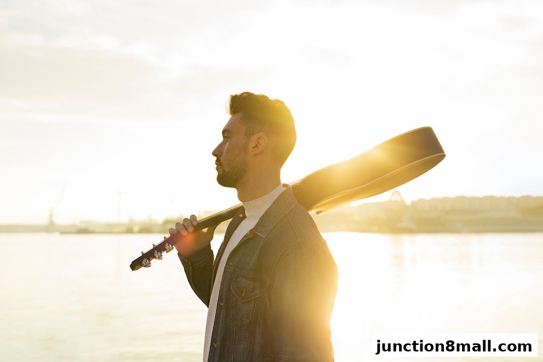 A man with a guitar case slung over his shoulder, looking out at the water during sunset.