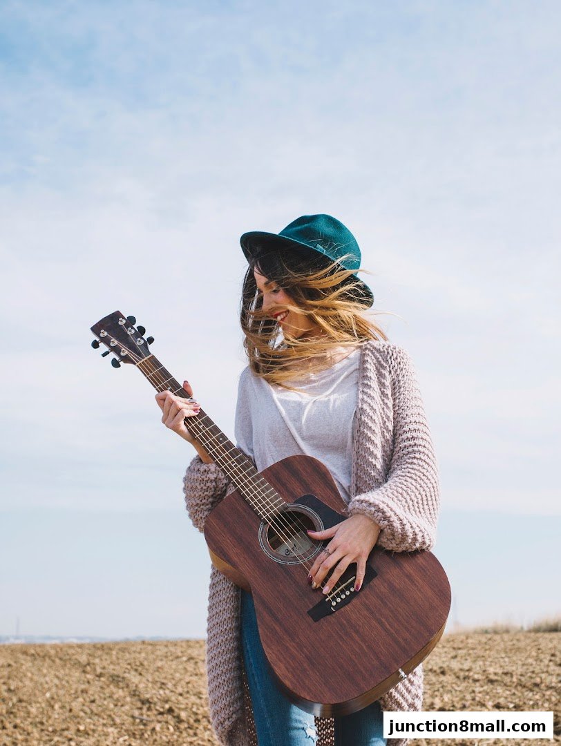 A smiling woman in a hat and cardigan plays an acoustic guitar outdoors.