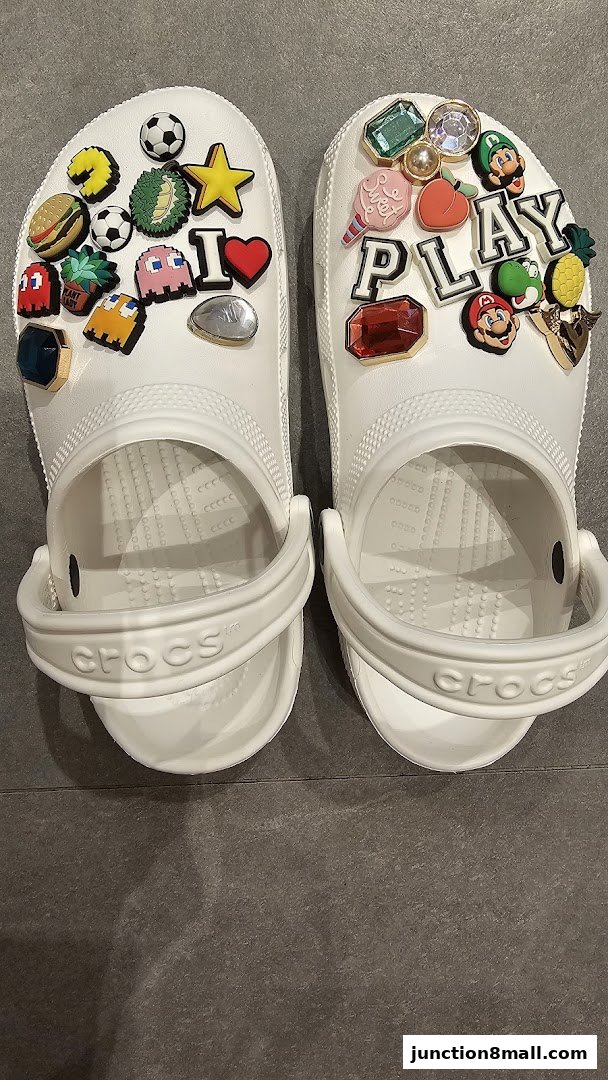 A pair of white Crocs adorned with various colorful Jibbitz charms, including food, gaming, and gemstone designs.
