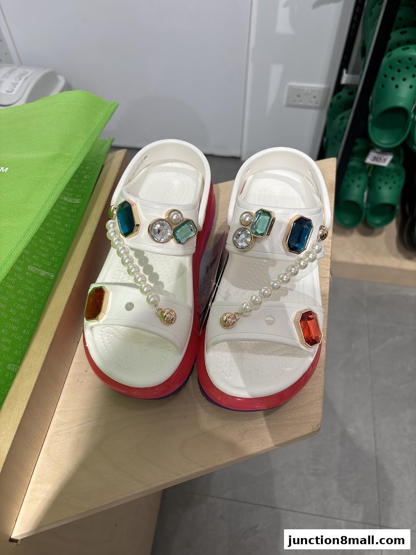 White Crocs sandals adorned with pearls and colorful gemstones displayed on a wooden surface.