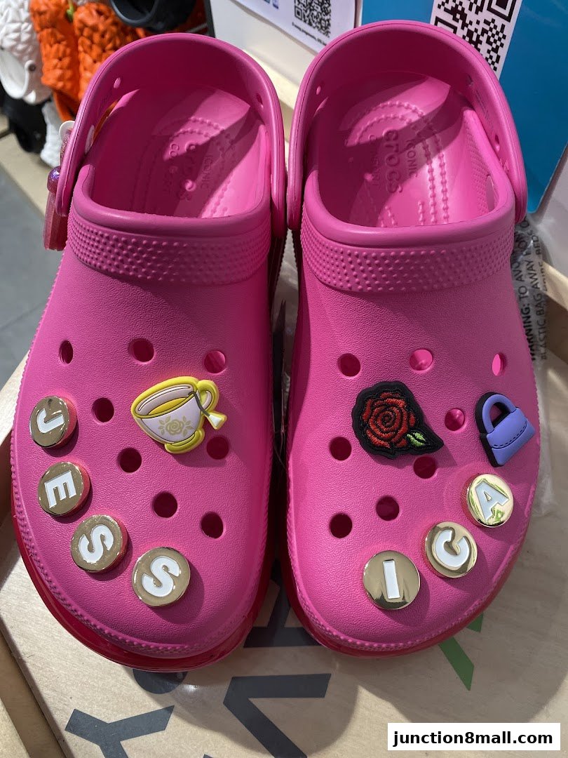 A pair of bright pink Crocs adorned with Jibbitz charms, including letters and decorative icons.