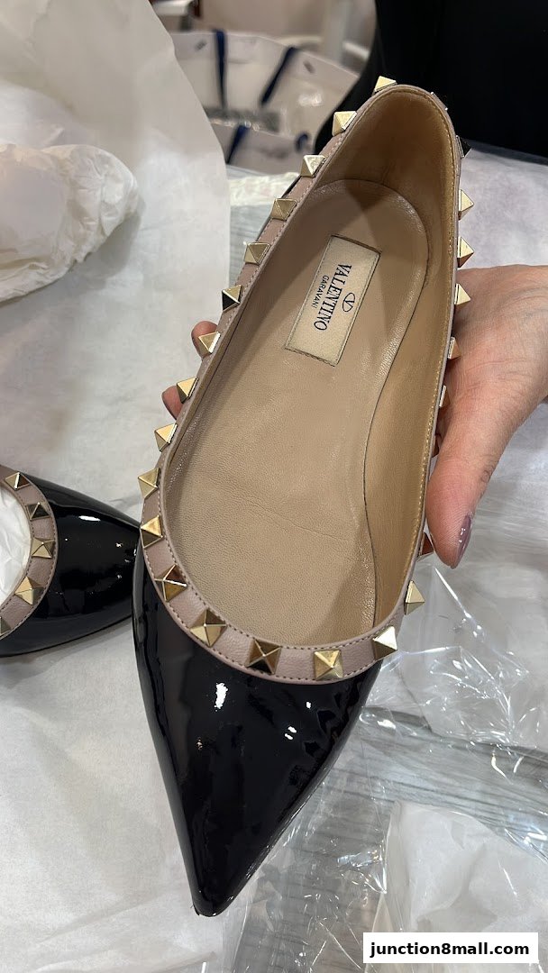 A hand holds a black patent leather Valentino shoe with gold studs.