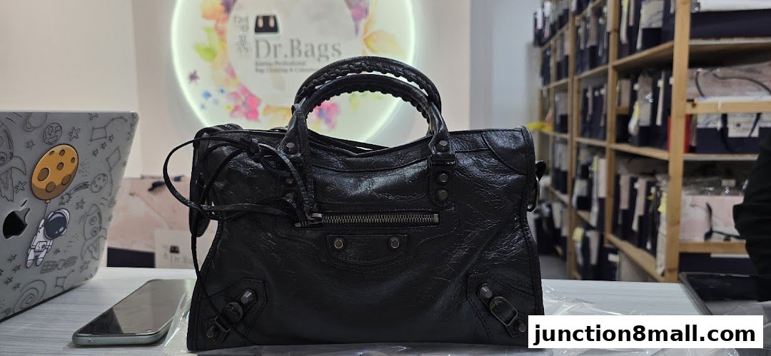 A black designer handbag is displayed on a table with a laptop and phone, with shelves of bags in the background.