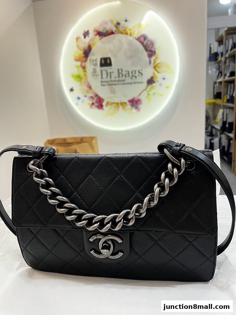 A black quilted handbag with a silver chain strap is displayed in front of a circular logo.