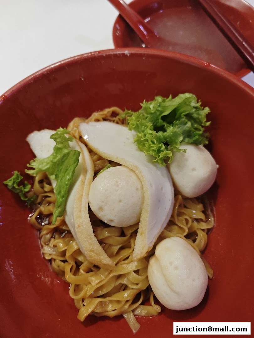 A close-up of a bowl of noodles with fish balls and lettuce, served in a red bowl.