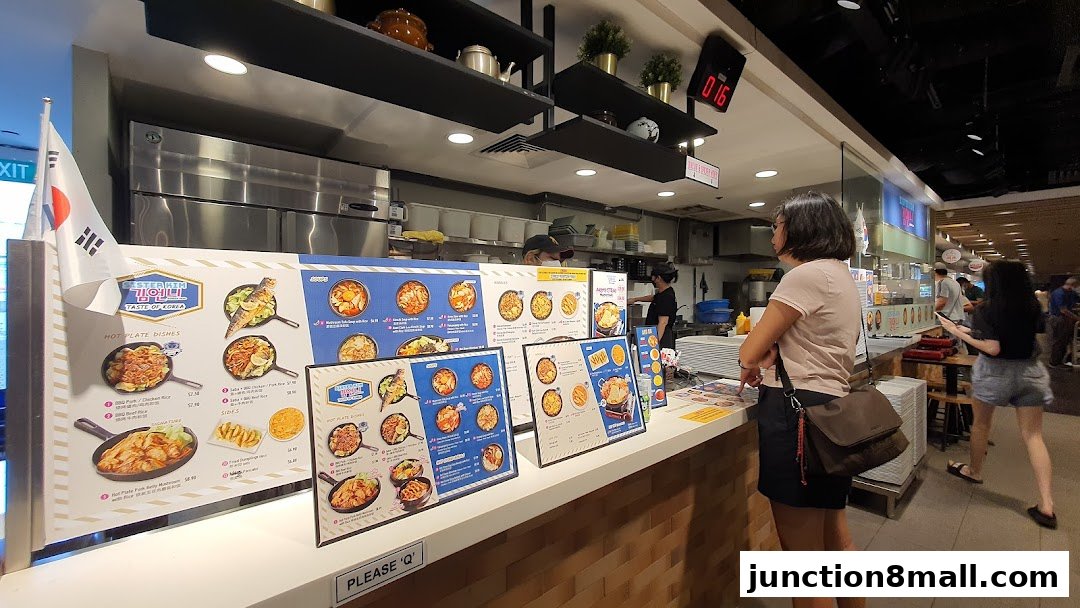 A food stall counter with Korean dishes displayed on menus and customers ordering.