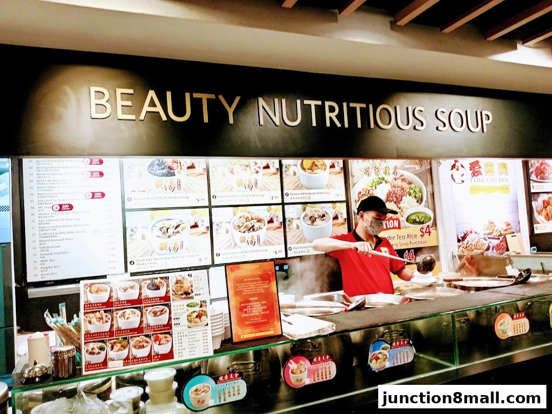A food stall counter with a menu board displaying various soups and a staff member serving food.