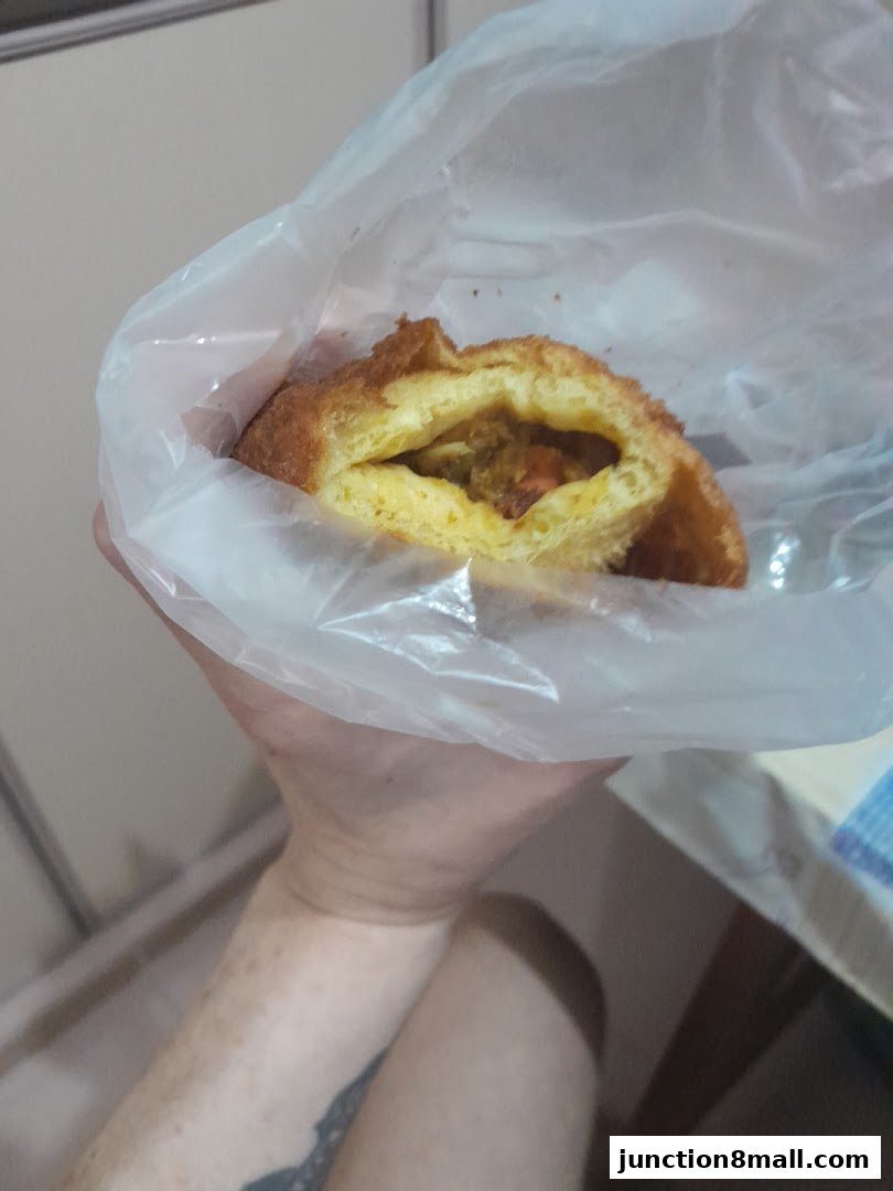 A hand holds a pastry filled with savory ingredients, wrapped in plastic.
