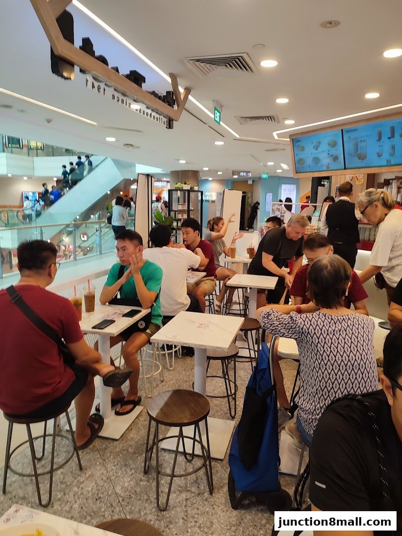 People dining at tables inside a bright, modern cafe with a view of a mall atrium.