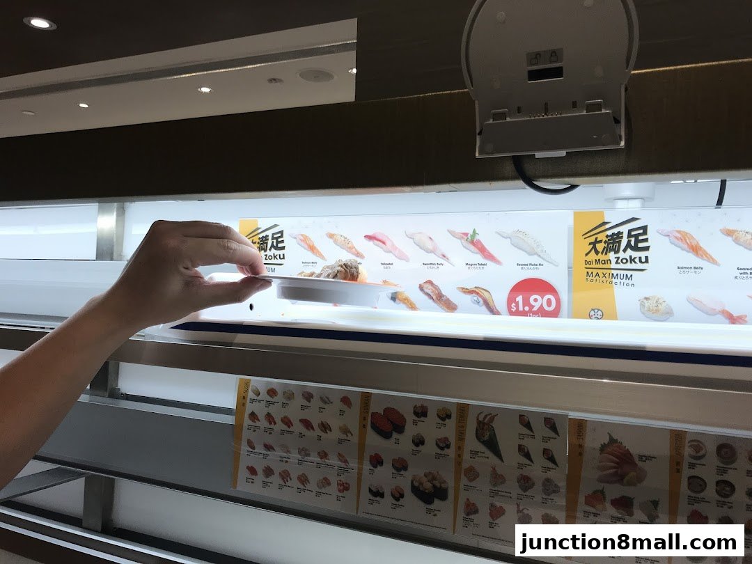 A hand reaches for a plate of sushi on a conveyor belt with a menu display.