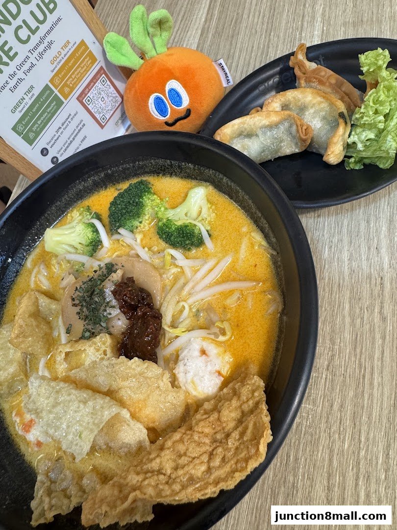 A bowl of Laksa with fried dumplings and lettuce, next to a promotional sign and a plush toy.