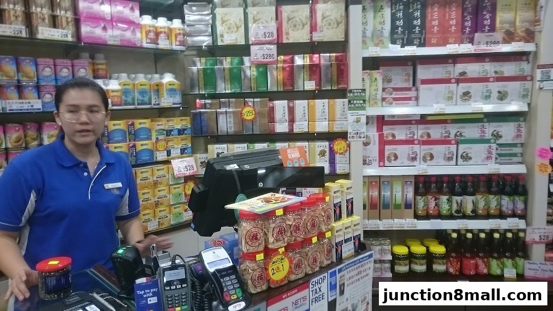 A shop assistant stands behind a counter filled with various health tonics and food products.