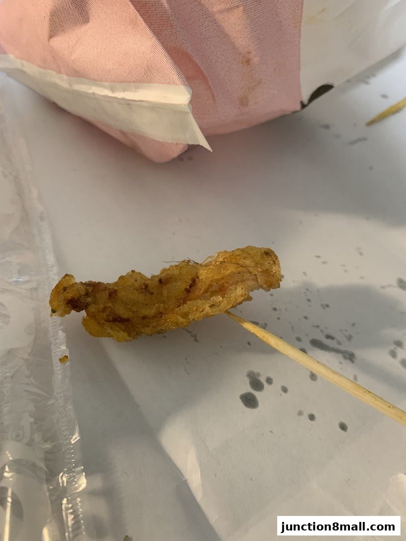 A close-up of a piece of fried chicken on a skewer, with a pink bag in the background.