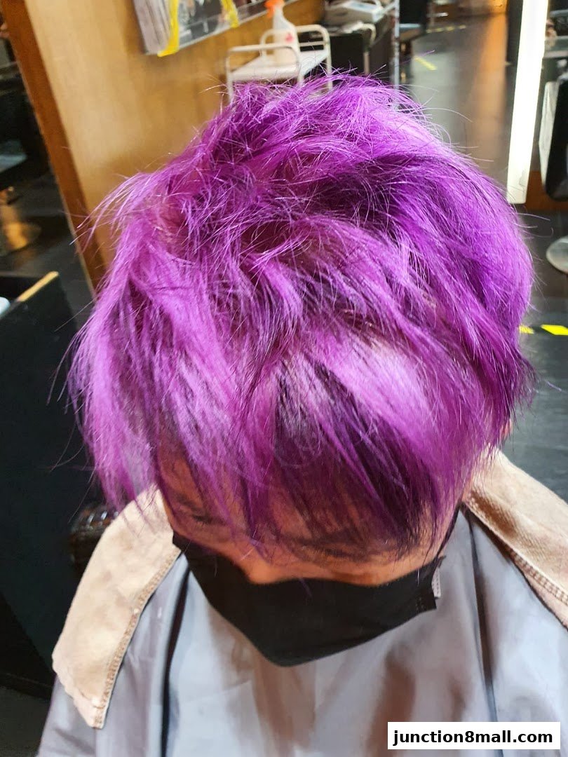 A person with vibrant purple hair wearing a black mask and a salon cape.