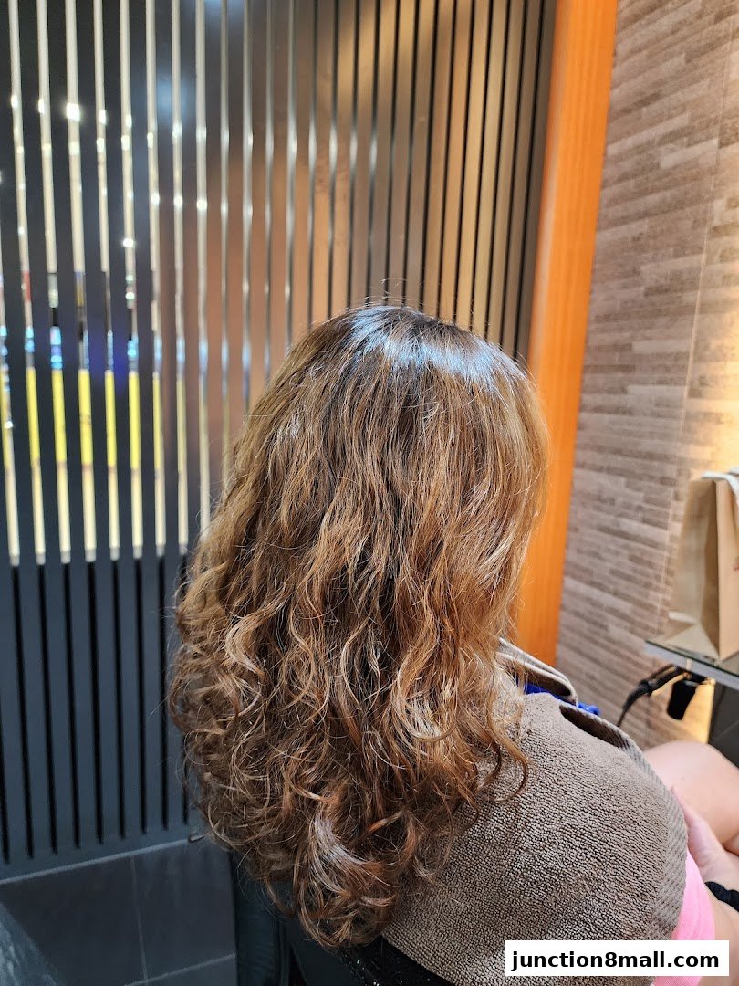 A person with curly, highlighted hair sits in a salon chair, draped in a towel.