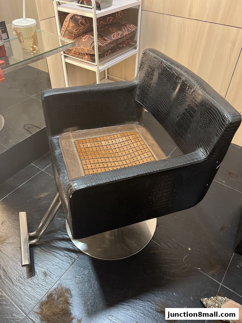 A black salon chair with a bamboo seat cushion sits on a tiled floor with scattered hair.
