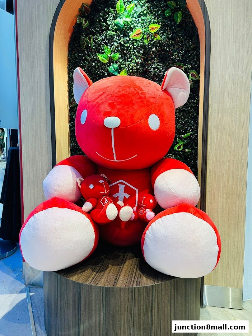 A large red teddy bear with smaller teddy bears sits in front of a green wall with plants.