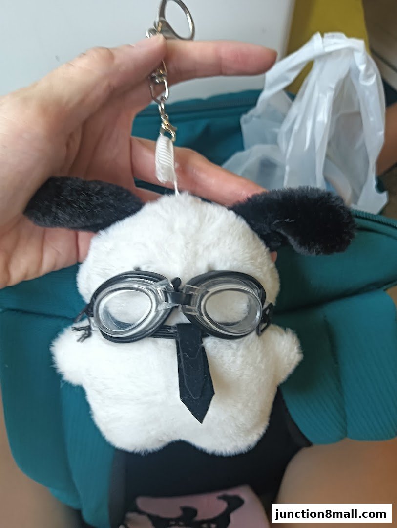 A hand holds a plush toy dog wearing goggles and a tie, attached to a keychain.