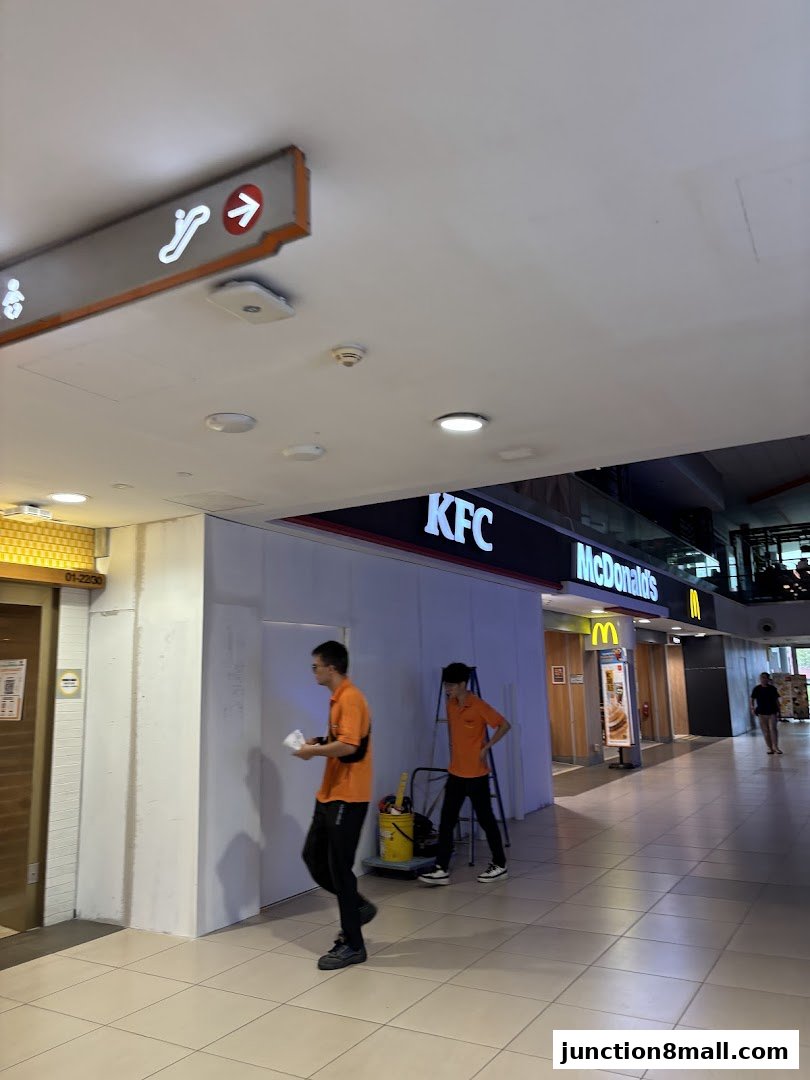 Two workers in orange shirts are near a KFC and McDonald's storefront.