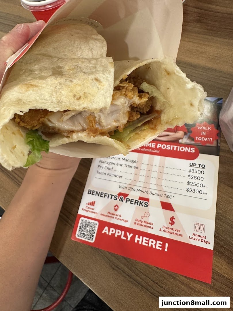 A hand holds a KFC chicken wrap with lettuce and sauce, next to a recruitment flyer.