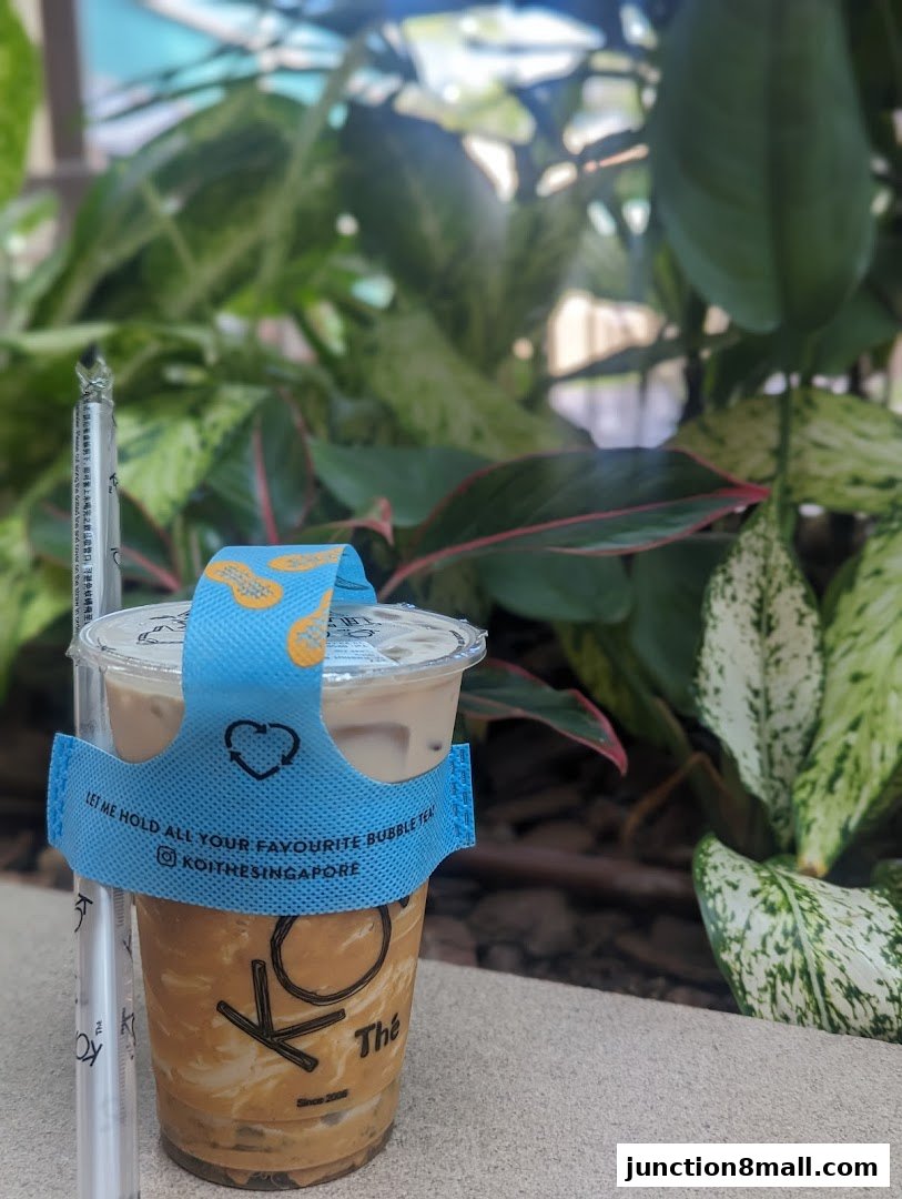 A cup of KOI Thé bubble tea with a blue holder and a straw, set against a backdrop of lush greenery.