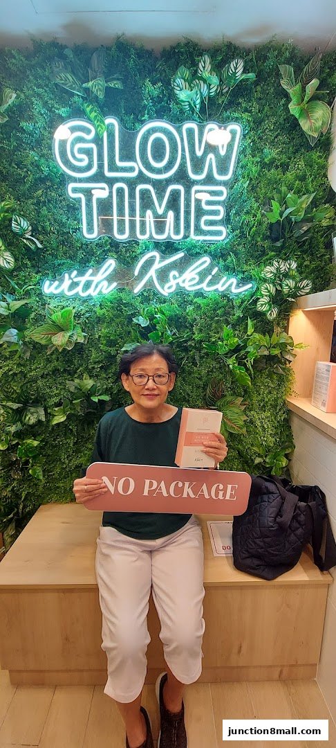 A woman sits in front of a green wall with a neon sign that says 'GLOW TIME with Kskin'.