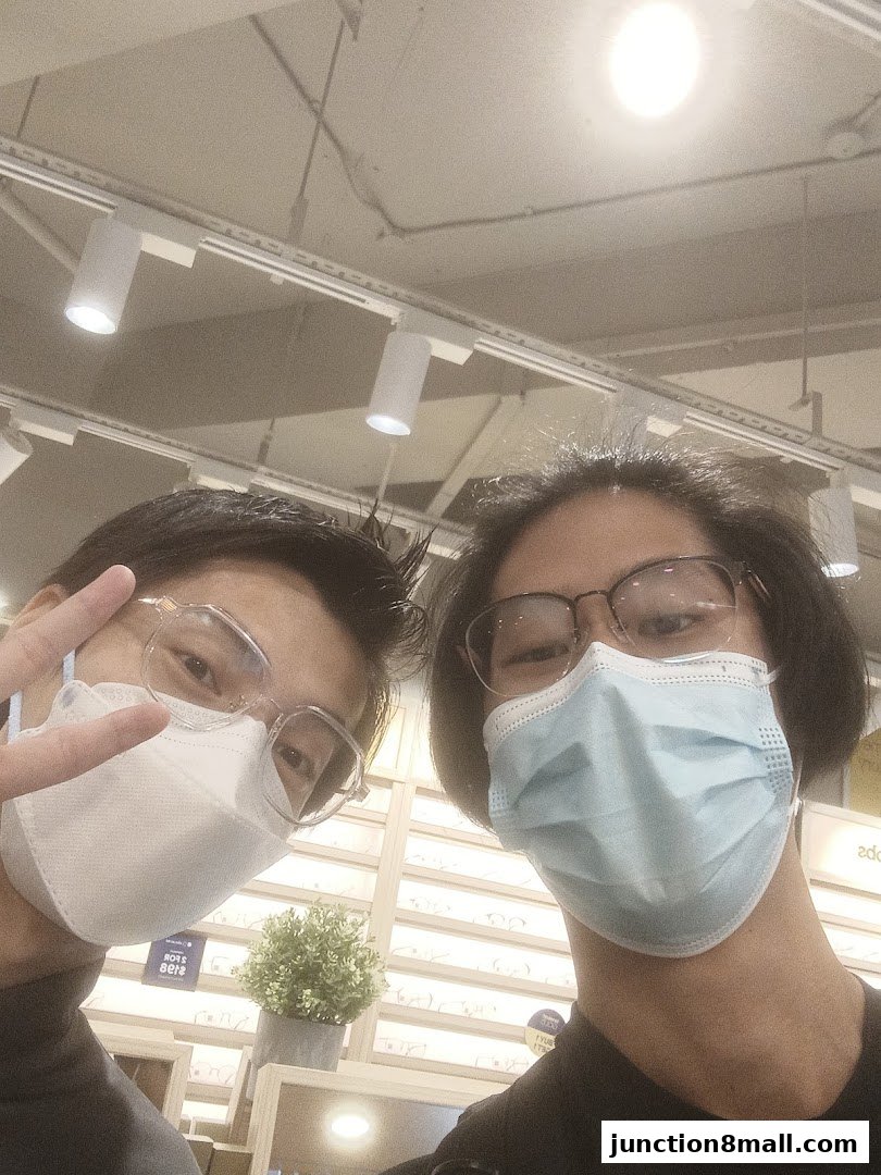 Two people wearing masks and glasses pose for a selfie in an optical shop.