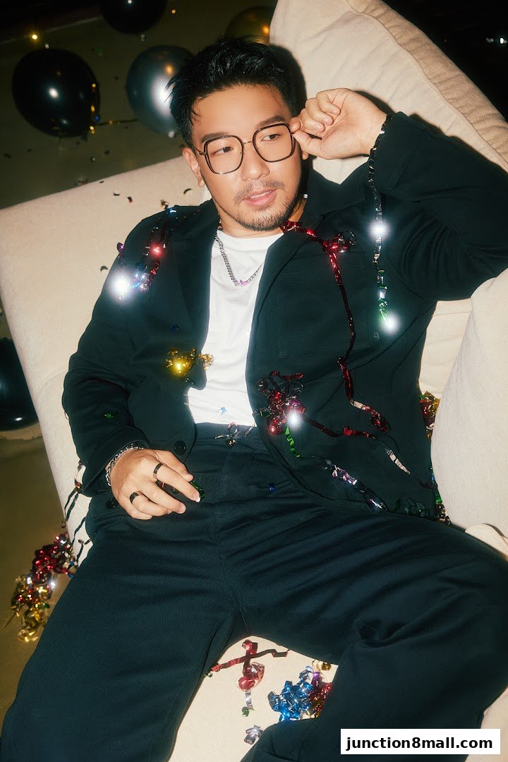 A man in a black suit with glasses sits on a couch surrounded by confetti and balloons.
