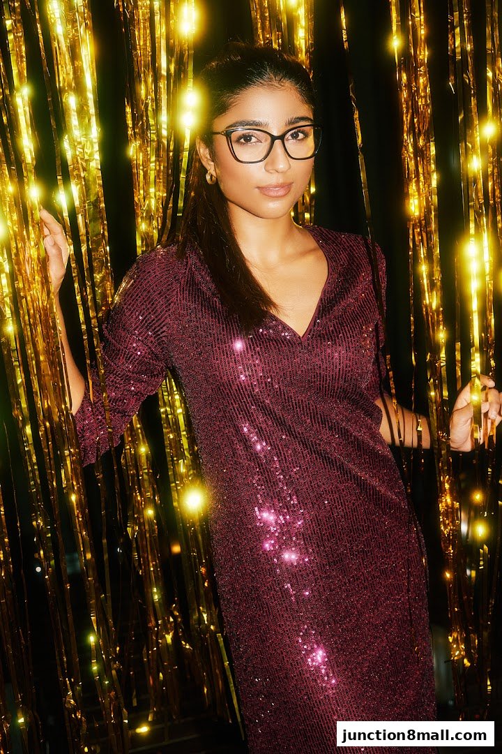 A woman wearing glasses and a sparkly dress poses against a backdrop of gold fringe.