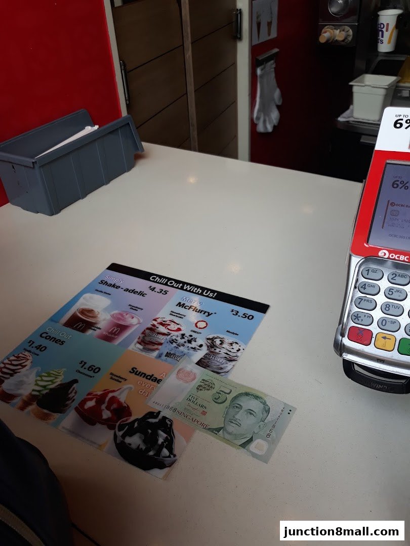 A McDonald's counter displaying a menu of shakes and McFlurrys, with a payment terminal and Singapore currency.