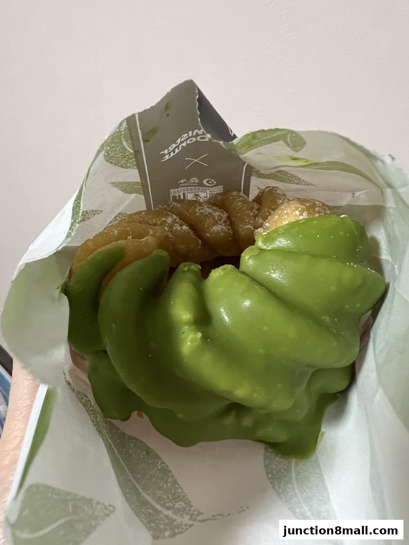 A close-up of a green-frosted donut with powdered sugar in a Mister Donut bag.