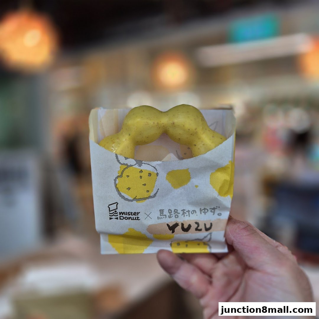 A hand holds a yellow Mister Donut with a yuzu flavor in a branded paper bag.