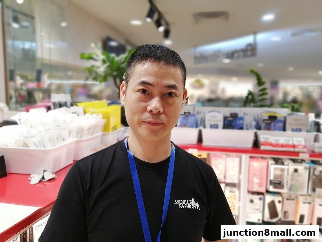 A man wearing a black t-shirt with the shop's logo stands in front of a display of phone accessories.