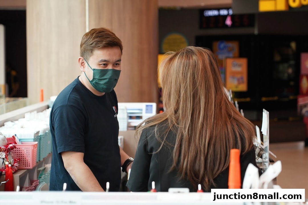 A masked employee speaks with a customer at a counter in a phone repair shop.