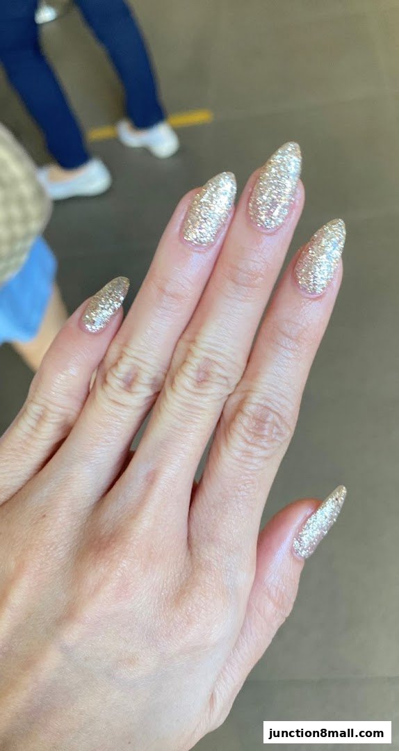 A close-up of a hand with long, almond-shaped nails covered in shimmering gold glitter.