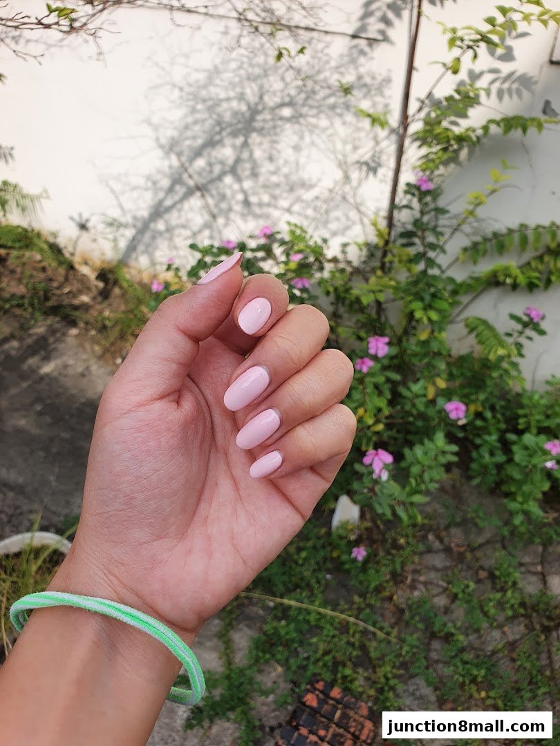 A hand with perfectly manicured light pink nails is shown against a backdrop of greenery and a textured wall.